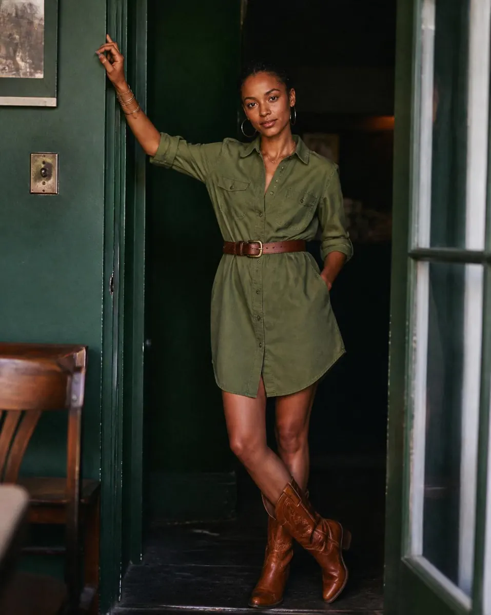 A petite woman in an olive belted shirt dress and tan boots in a green doorway